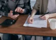 Two people working together on tax forms using a calculator at a wooden desk.
