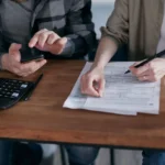 Two people working together on tax forms using a calculator at a wooden desk.