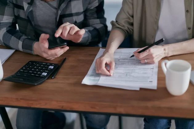 Two people working together on tax forms using a calculator at a wooden desk. Two people working together on tax forms using a calculator at a wooden desk.