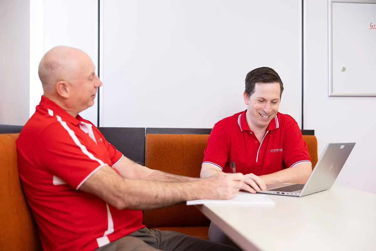 two men sitting on a couch in front of a white desk talking and making notes on a laptop and on paper two men sitting on a couch in front of a white desk talking and making notes on a laptop and on paper