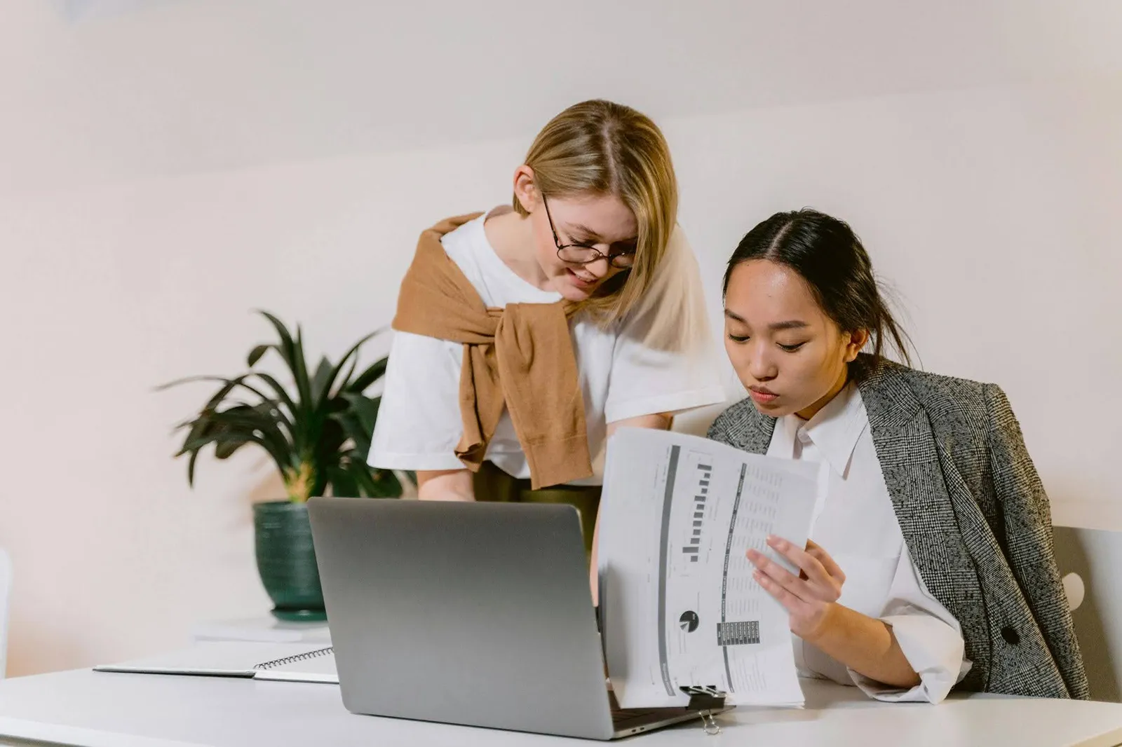 two females sitting at a desk looking at a document and laptop