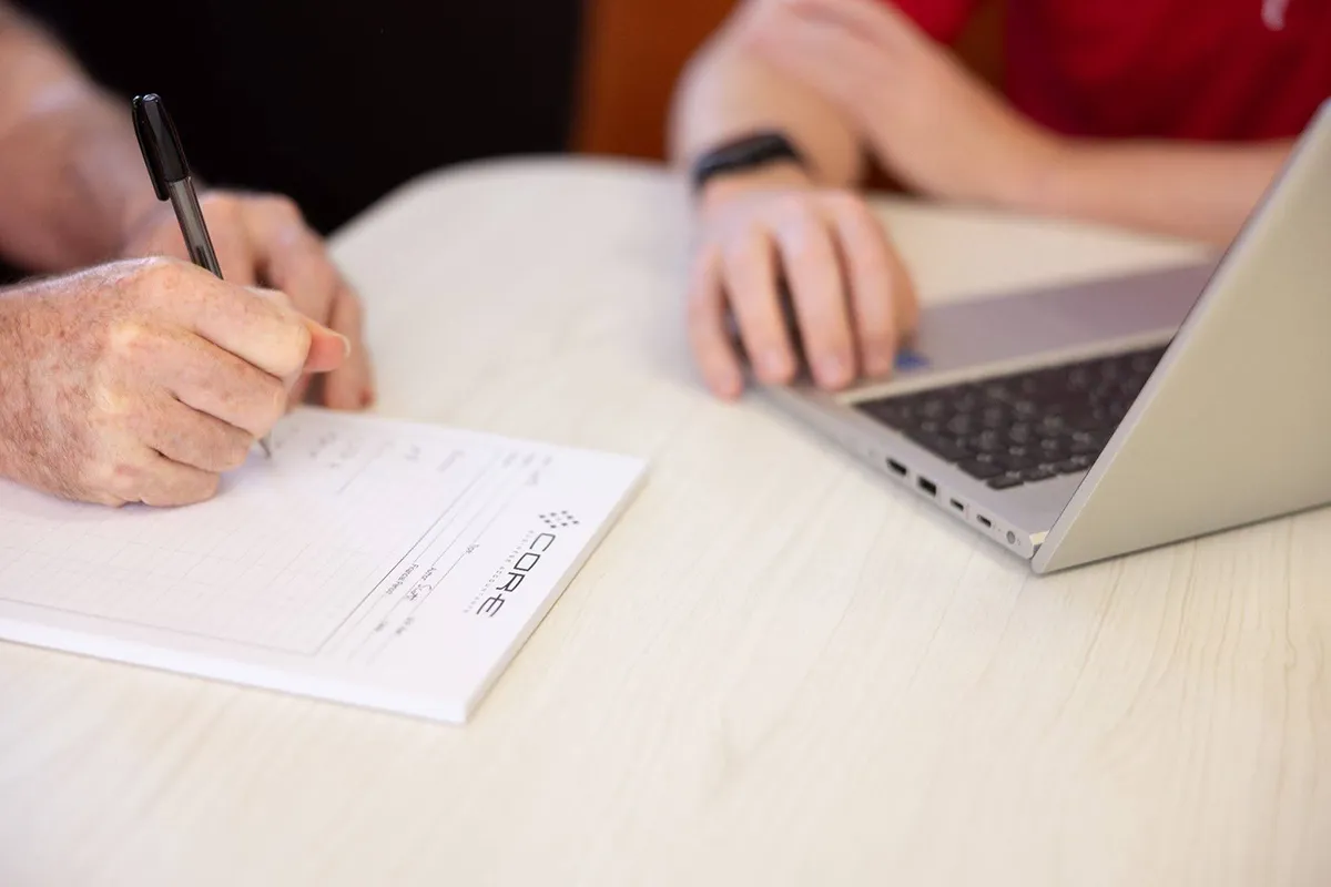 two people writing on a notepad and using a laptop two people writing on a notepad and using a laptop
